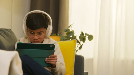 Kleiner-Junge-Sitzt-Zu-Hause-Auf-Dem-Sofa,-Trägt-Kabellose-Kopfhörer-Und-Spielt-Spiele-Oder-Streamt-Auf-Ein-Digitales-Tablet